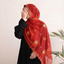 A woman wearing a black abaya and a bright red embroidered hijab with gold floral patterns and scalloped edges, standing against a light beige wall with green leaves at the side.