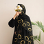 A woman wearing a black hijab and long black dress with gold embroidered geometric patterns, holding white flowers in front of her face against a plain light beige wall with a green plant in the background.
