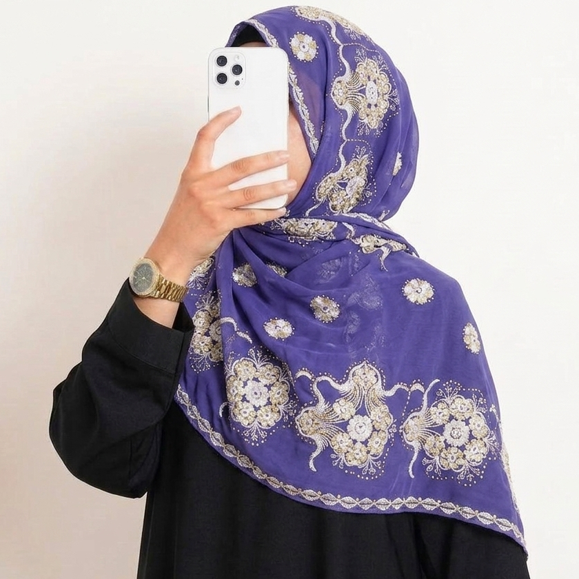 A person taking a mirror selfie wearing a purple hijab with a white floral pattern and a black long-sleeve top against a plain off-white wall.