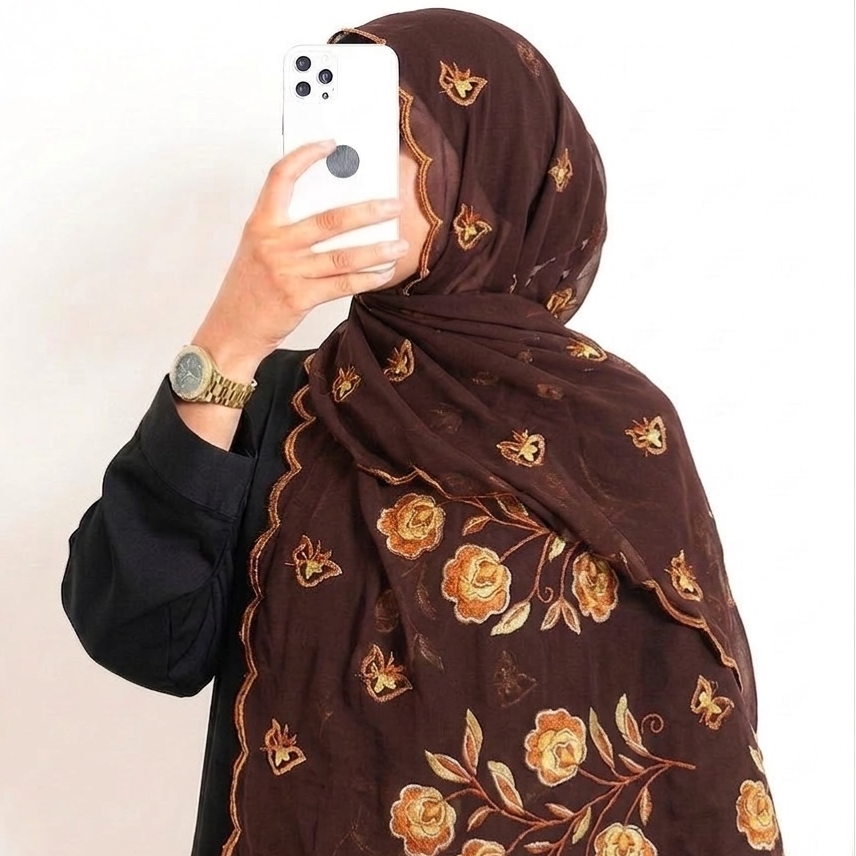 A person taking a mirror selfie wearing a dark brown hijab with embroidered gold and beige floral motifs and a scalloped edge, paired with a black top. A white smartphone is held up to take the photo.