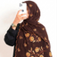 A person taking a mirror selfie wearing a dark brown hijab with embroidered gold and beige floral motifs and a scalloped edge, paired with a black top. A white smartphone is held up to take the photo.