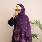 A woman wearing a deep purple hijab with embroidered floral patterns and a black dress, holding a bouquet of white flowers against her face. A green plant is visible on the right side of the background.