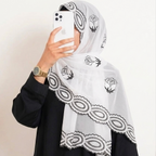 A woman wearing a white hijab with black embroidered rose motifs and scalloped lace trim, paired with a black outfit. She is holding a white smartphone to take a mirror selfie against a plain off-white wall.