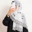 A woman wearing a white hijab with black embroidered rose motifs and scalloped lace trim, paired with a black outfit. She is holding a white smartphone to take a mirror selfie against a plain off-white wall.