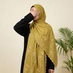 A woman wearing a black abaya and a mustard yellow embroidered hijab, standing against a beige wall with potted plants on either side.