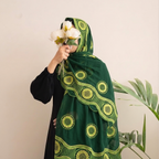 A woman wearing a black outfit and a dark green embroidered hijab with yellow-green circular patterns and scalloped borders, holding white flowers against a light beige wall with a green plant in the background.