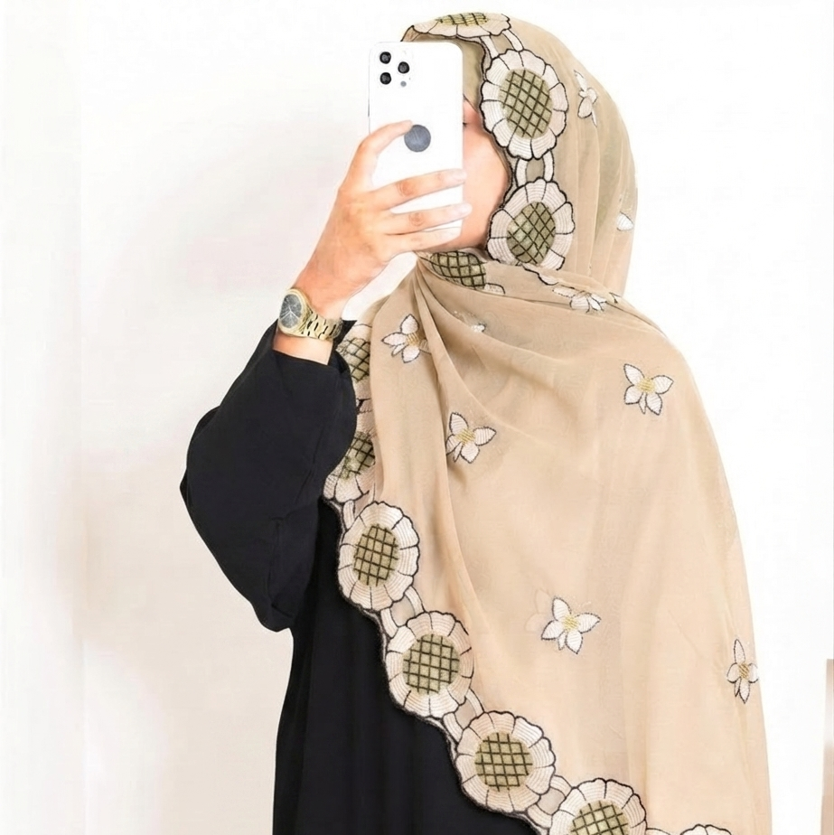 A woman wearing a beige hijab with embroidered floral and butterfly patterns, paired with a black abaya, taking a mirror selfie with a white smartphone.