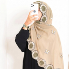 A woman wearing a beige hijab with embroidered floral and butterfly patterns, paired with a black abaya, taking a mirror selfie with a white smartphone.