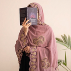 A woman wearing a dusty pink hijab with gold and purple embroidered patterns and a scalloped border, holding a book titled 'Prayers Book' in front of her face. She is wearing a black dress, standing against a beige wall with a green plant in the background.