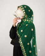 A woman wearing a dark green sheer hijab with gold coin embroidery and a scalloped border, paired with a black abaya, holding a large white flower in front of her face against a light gray background.